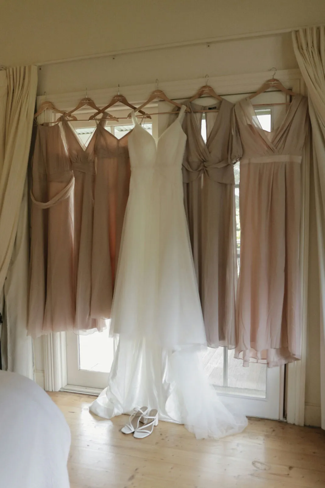 Wedding dress and bridesmaid dresses in soft blush and mauve hanging together in a sunlit room