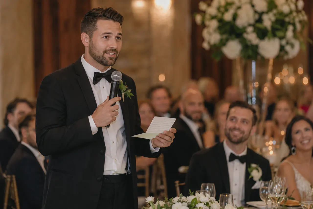 Brother of the groom giving a wedding toast at the reception while guests look on smiling