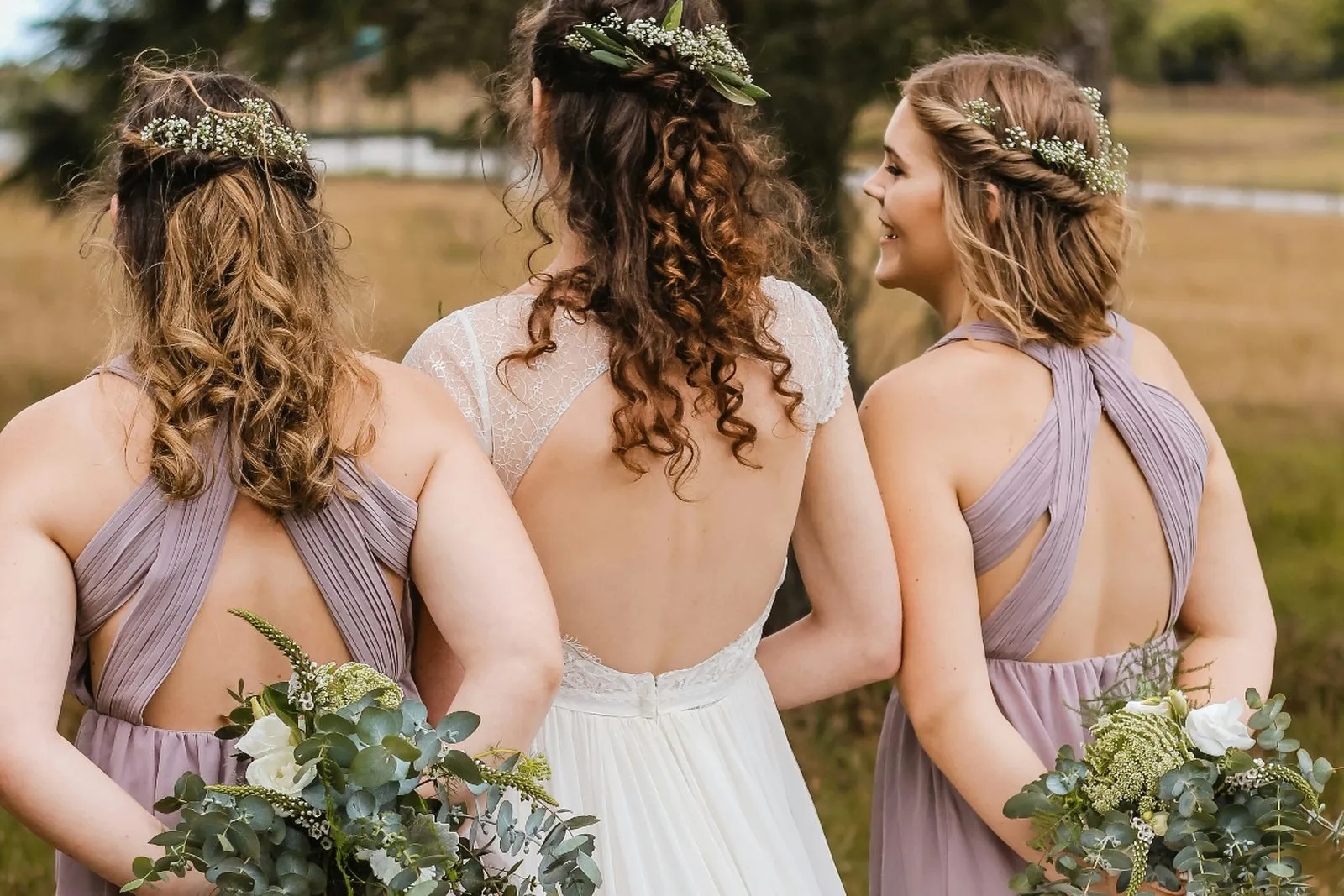 Bride with two bridesmaids in lilac dresses holding eucalyptus bouquets in a sunlit field