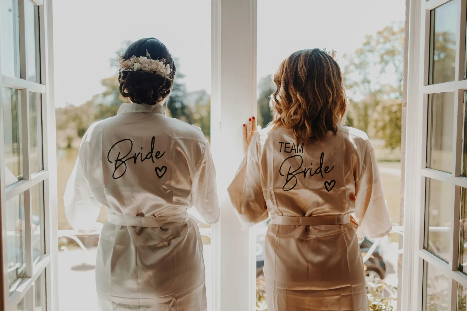 Bride and a bridesmaid wearing 'Bride' and 'Team Bride' robes looking out an open window together
