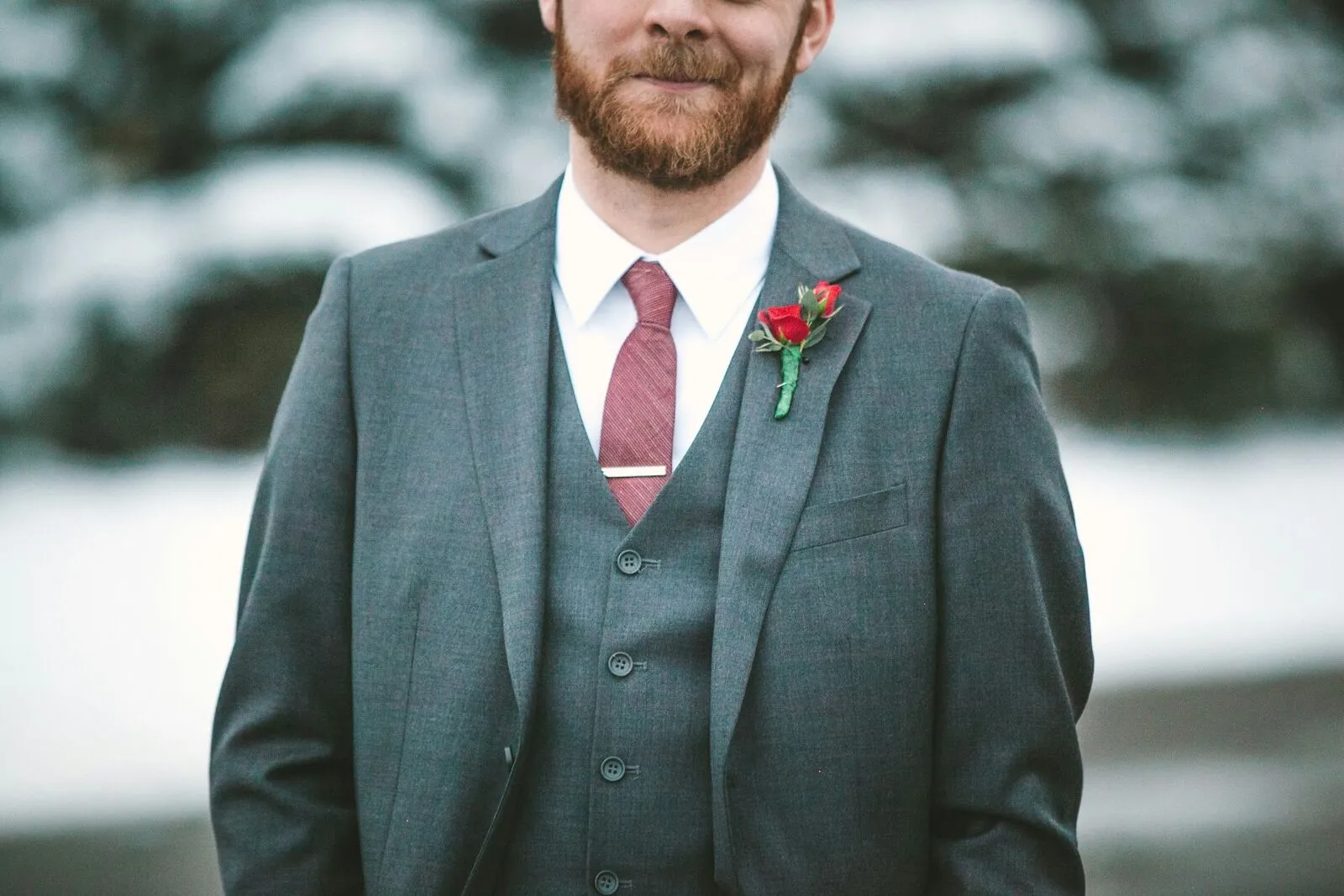 Groom in a grey three-piece suit with a red tie and rose boutonniere standing in a winter landscape