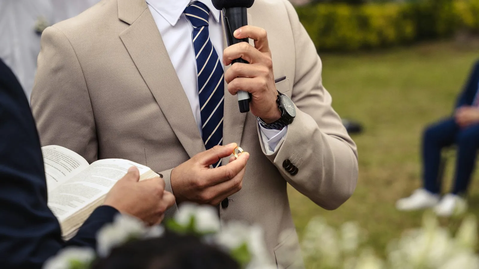 Groom in a beige suit holding a microphone and a wedding ring during his speech