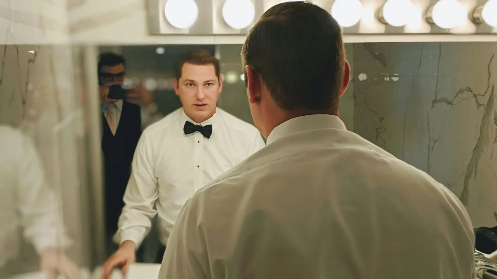 Groom in a white tuxedo shirt and bow tie practicing his speech in the mirror before the wedding
