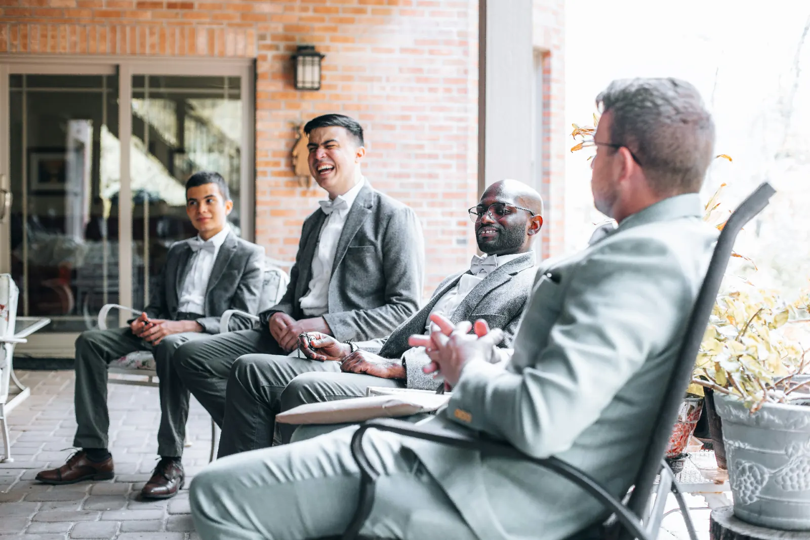 Groomsmen sitting together on a sunlit patio, one laughing as the others share a relaxed moment before the wedding