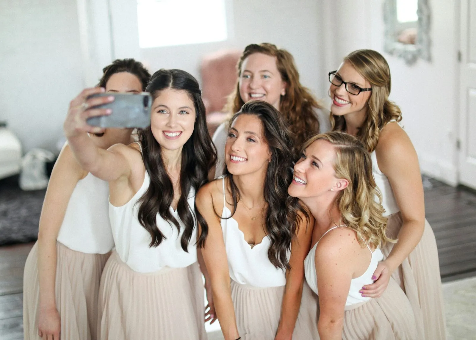 Bridesmaids in soft white tops and blush skirts taking a smiling selfie together in a bright bridal suite