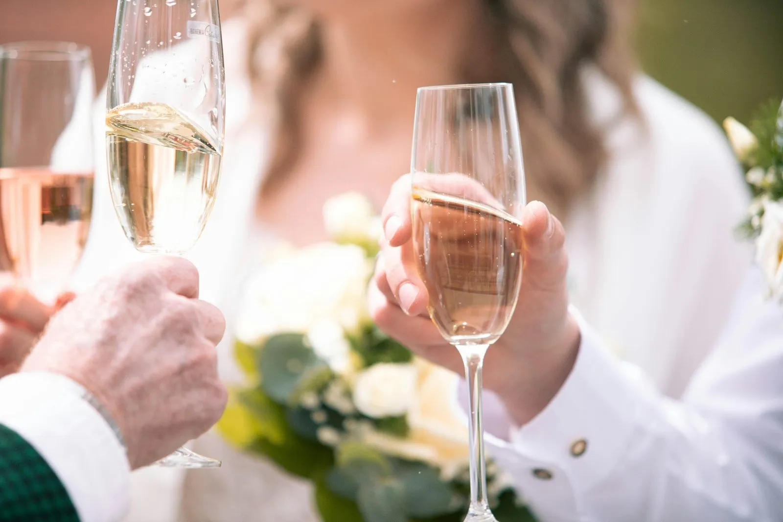 Hands clinking champagne flutes in a warm toast in front of a soft-focus wedding bouquet