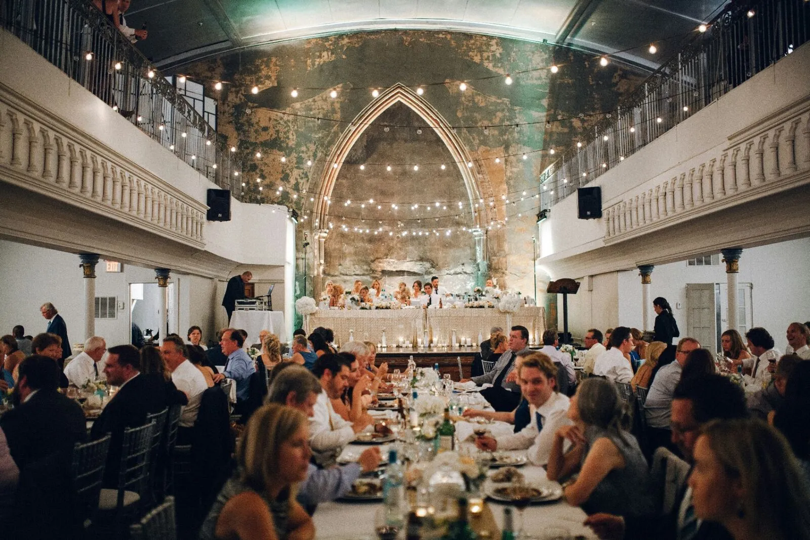 Wedding reception guests dining and laughing under string lights inside an elegant historic venue