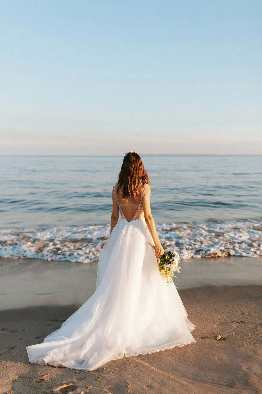 Bride in a flowing white gown standing barefoot on the sand looking out toward the sea at sunset