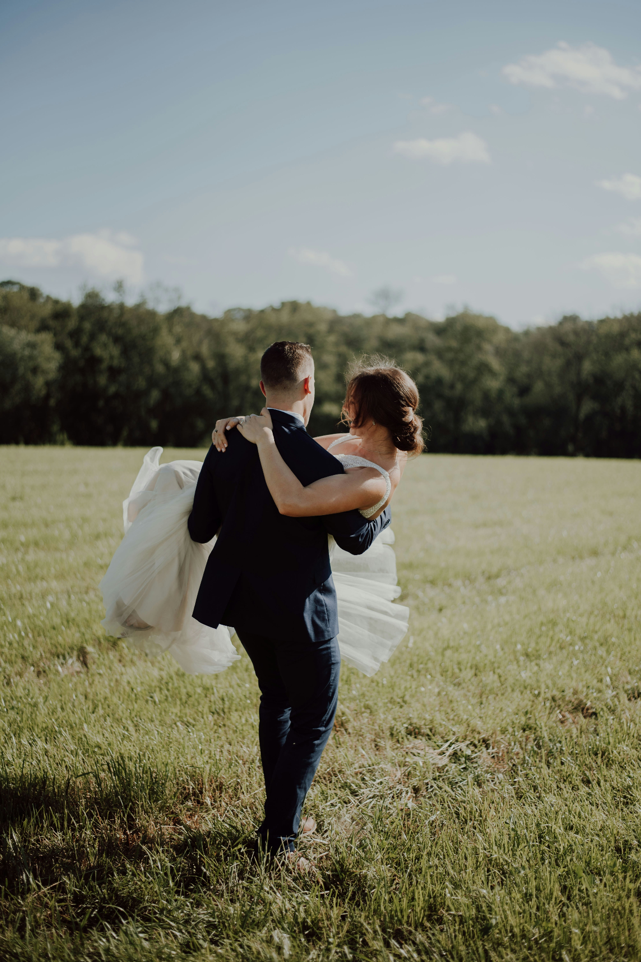 Groom carrying bride through a sunlit meadow on their wedding day