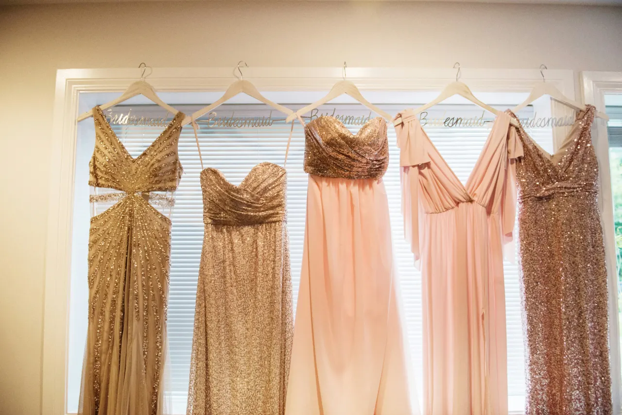 Bridesmaids dresses in blush, gold, and rose hanging on personalized hangers in front of a sunlit window