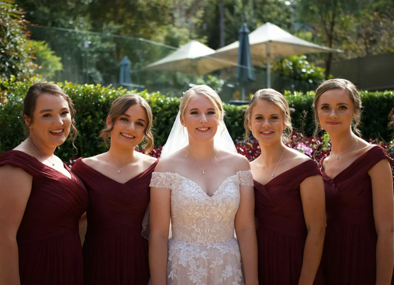 Bride in a lace gown standing with her four bridesmaids in burgundy dresses outside a garden venue