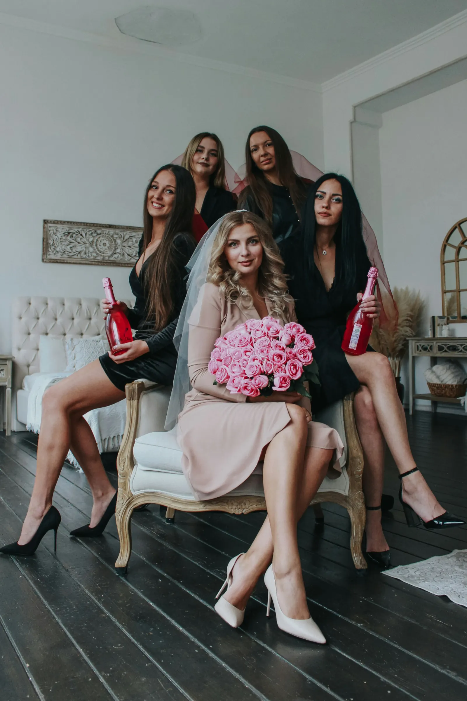 Bride seated with her bridesmaids in black, holding pink champagne and a bouquet of pink roses