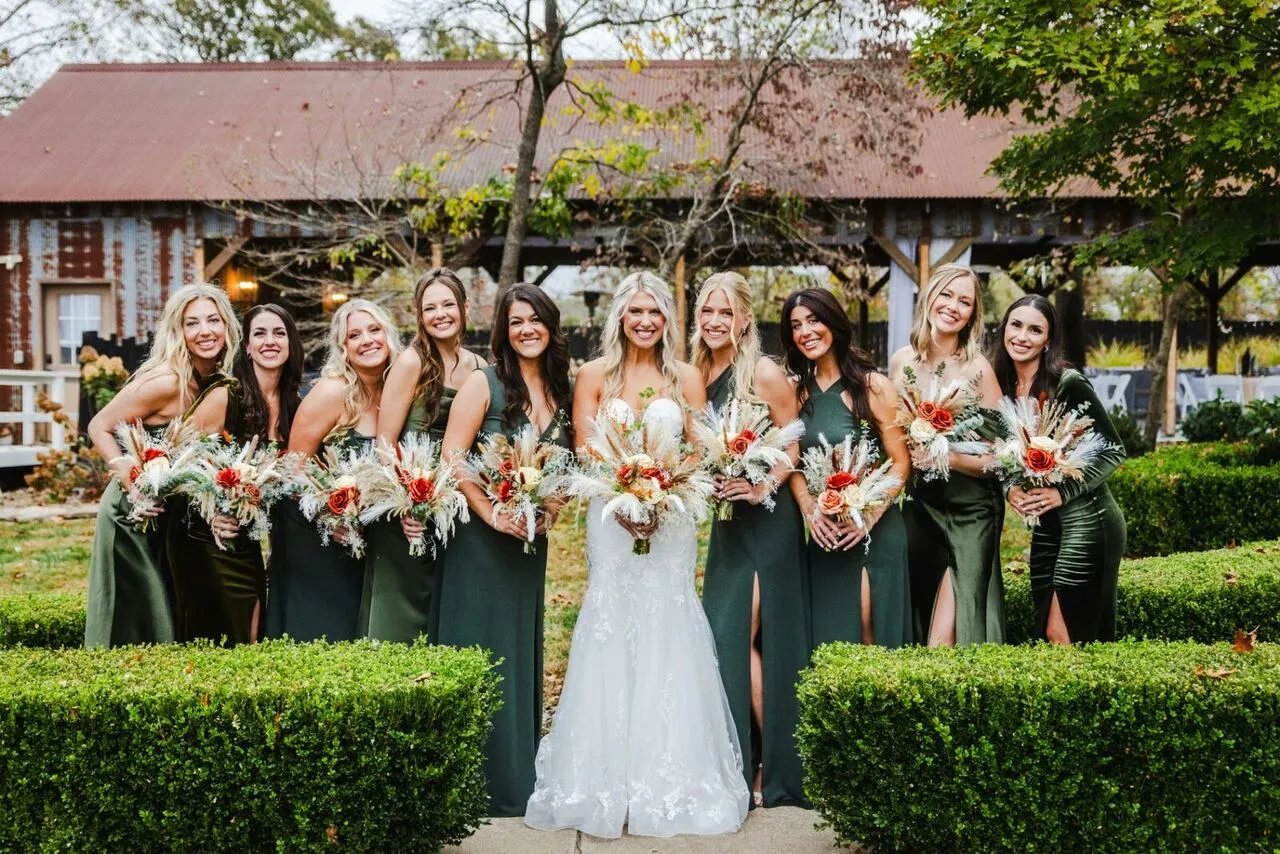 Bride and bridesmaids in deep green dresses smiling together outdoors with pampas and rose bouquets