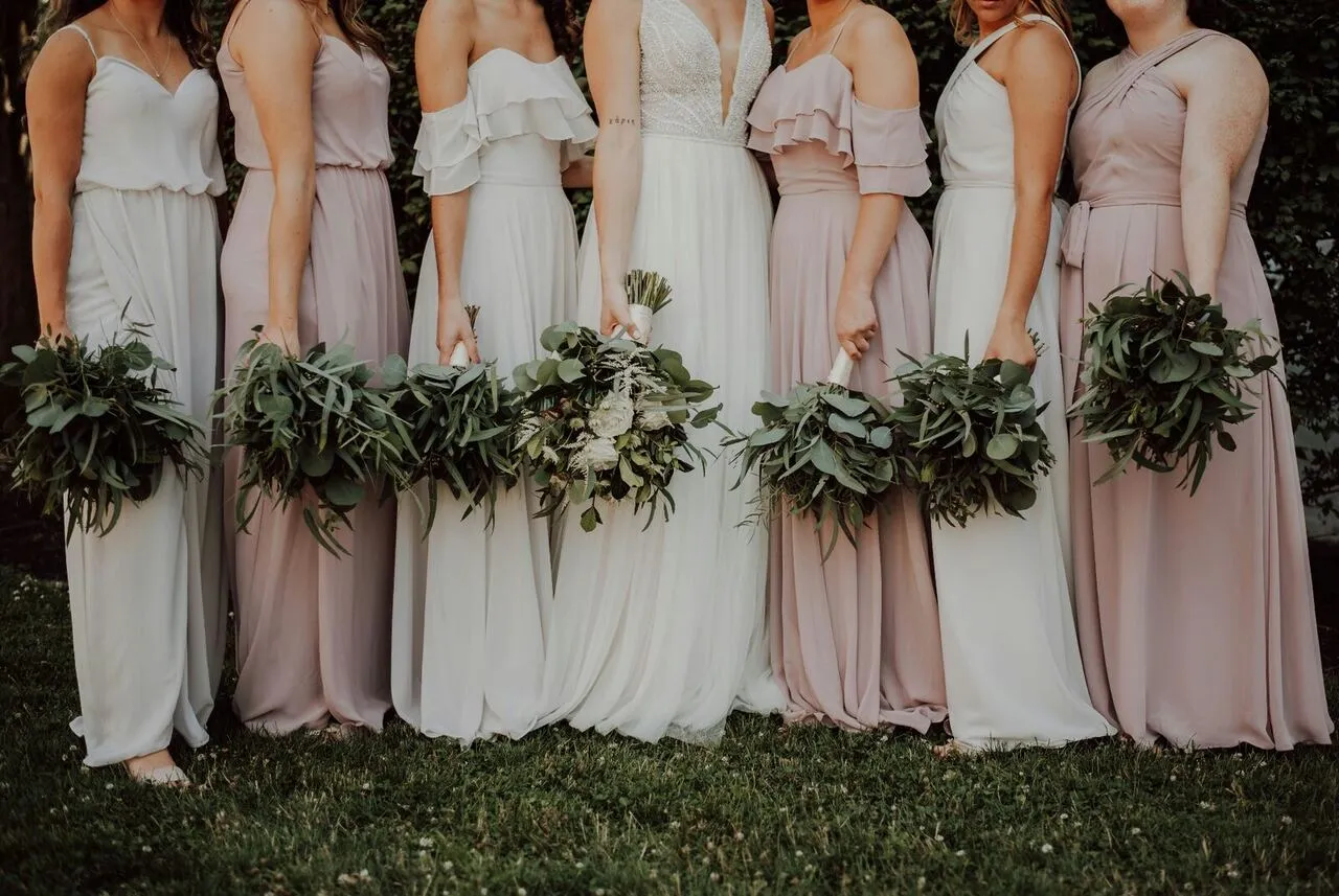 Bride and bridesmaids in soft white and blush gowns holding eucalyptus bouquets in a sunlit garden
