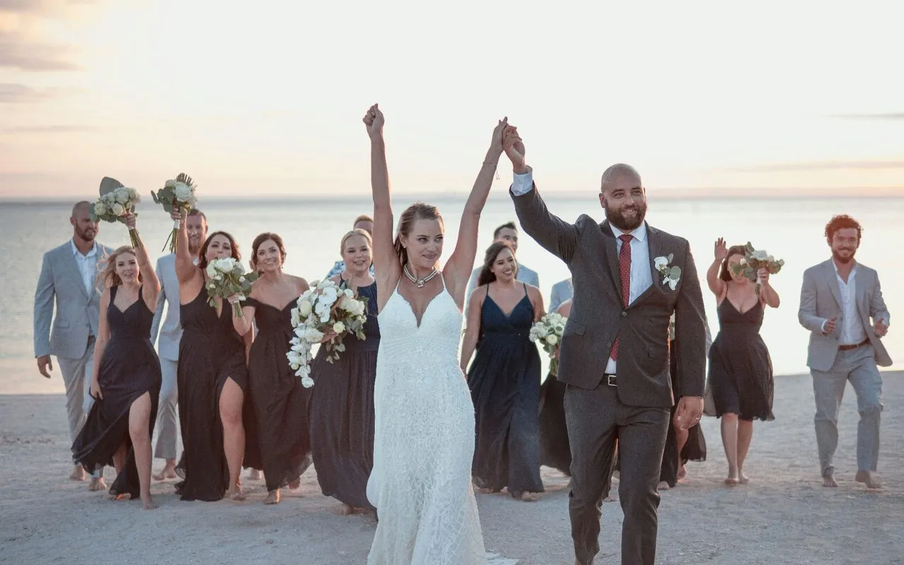 Bride and groom raising joined hands with the wedding party celebrating on a beach at sunset