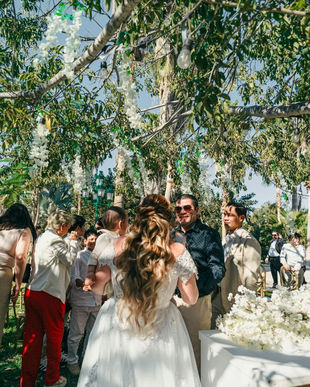 Bride in lace gown greeting guests under a canopy of trees and hanging white florals at her reception