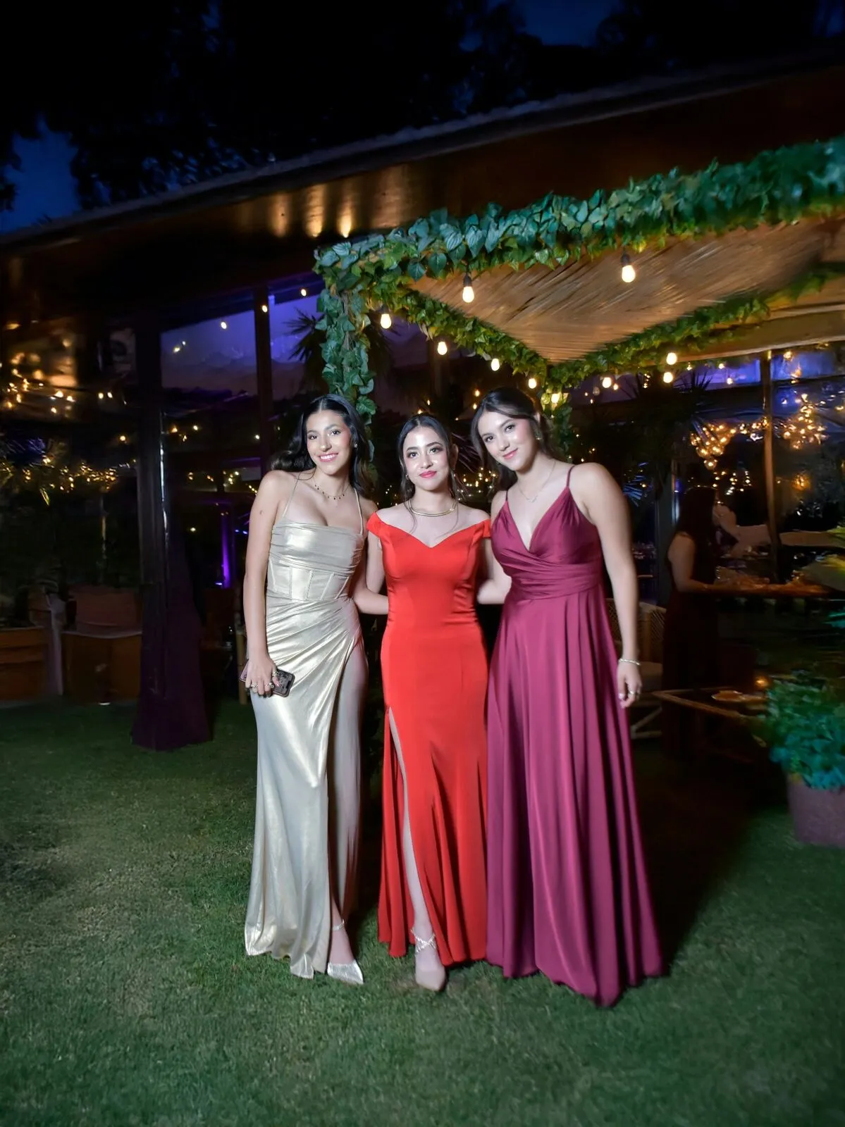 Three young women in elegant gold, red, and burgundy gowns smiling together at an evening reception