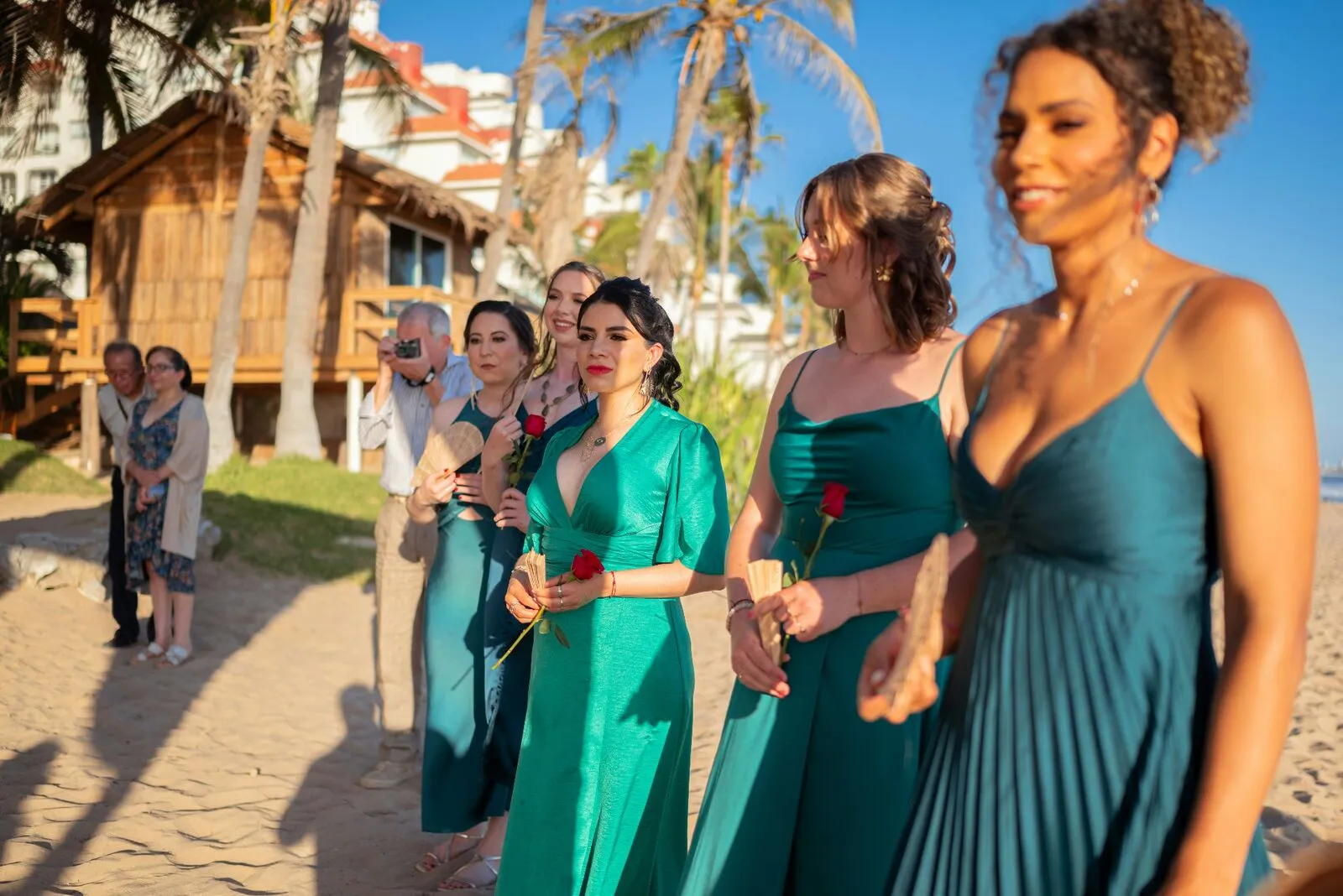 Bridesmaids in emerald satin gowns walking down a sunlit beach aisle holding red roses