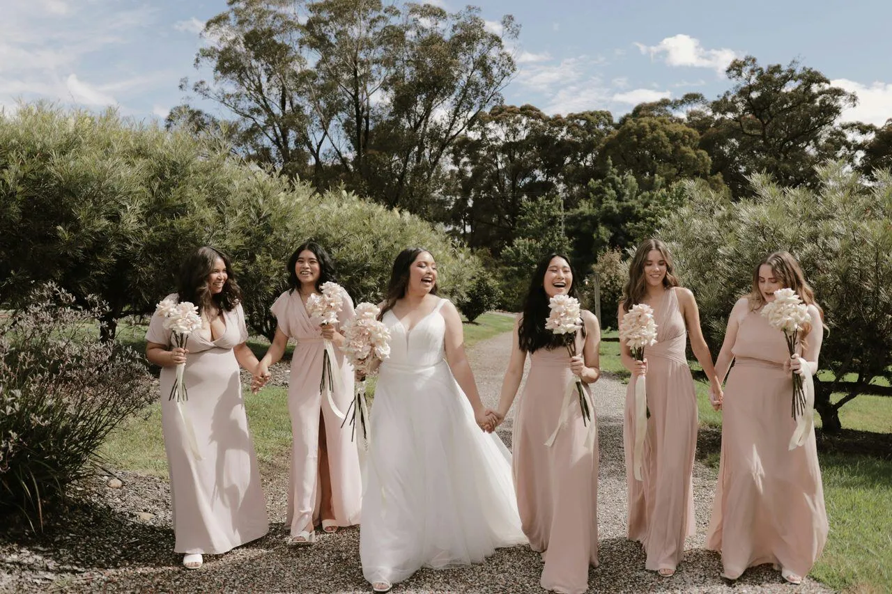 Bride and bridesmaids in blush gowns walking together holding bouquets on a garden path