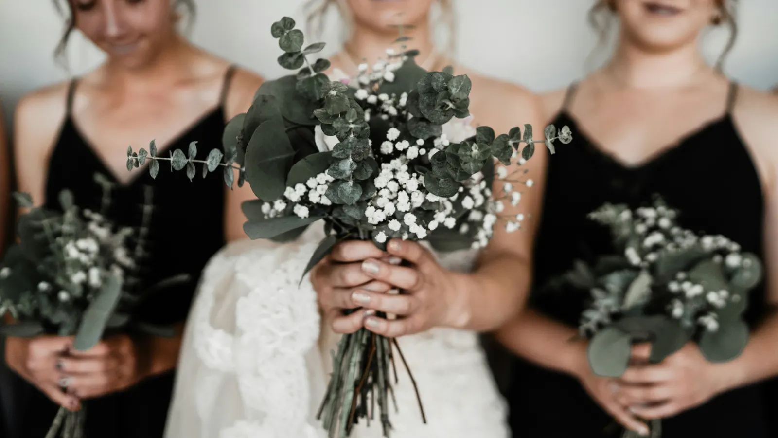 Bride holding a eucalyptus and baby's breath bouquet flanked by her sisters in black slip dresses with matching greenery bouquets