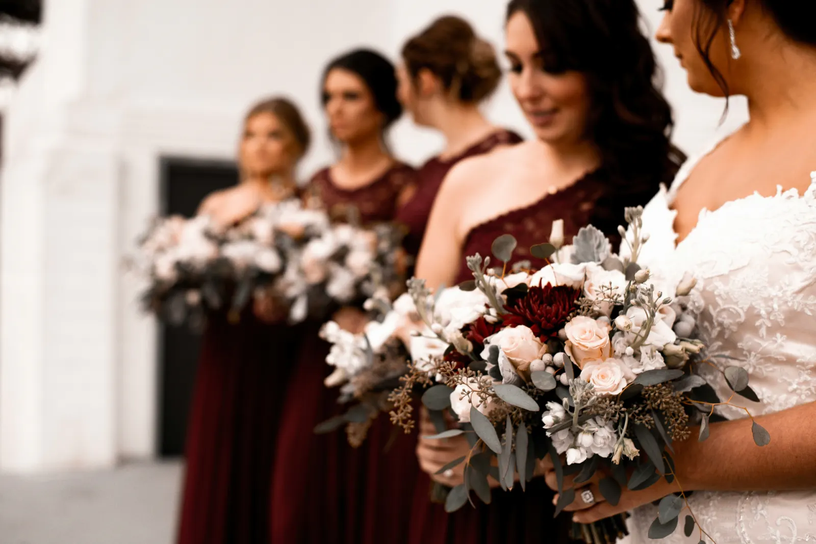Bride in lace beside her sisters and bridesmaids in burgundy gowns holding pale rose and eucalyptus bouquets