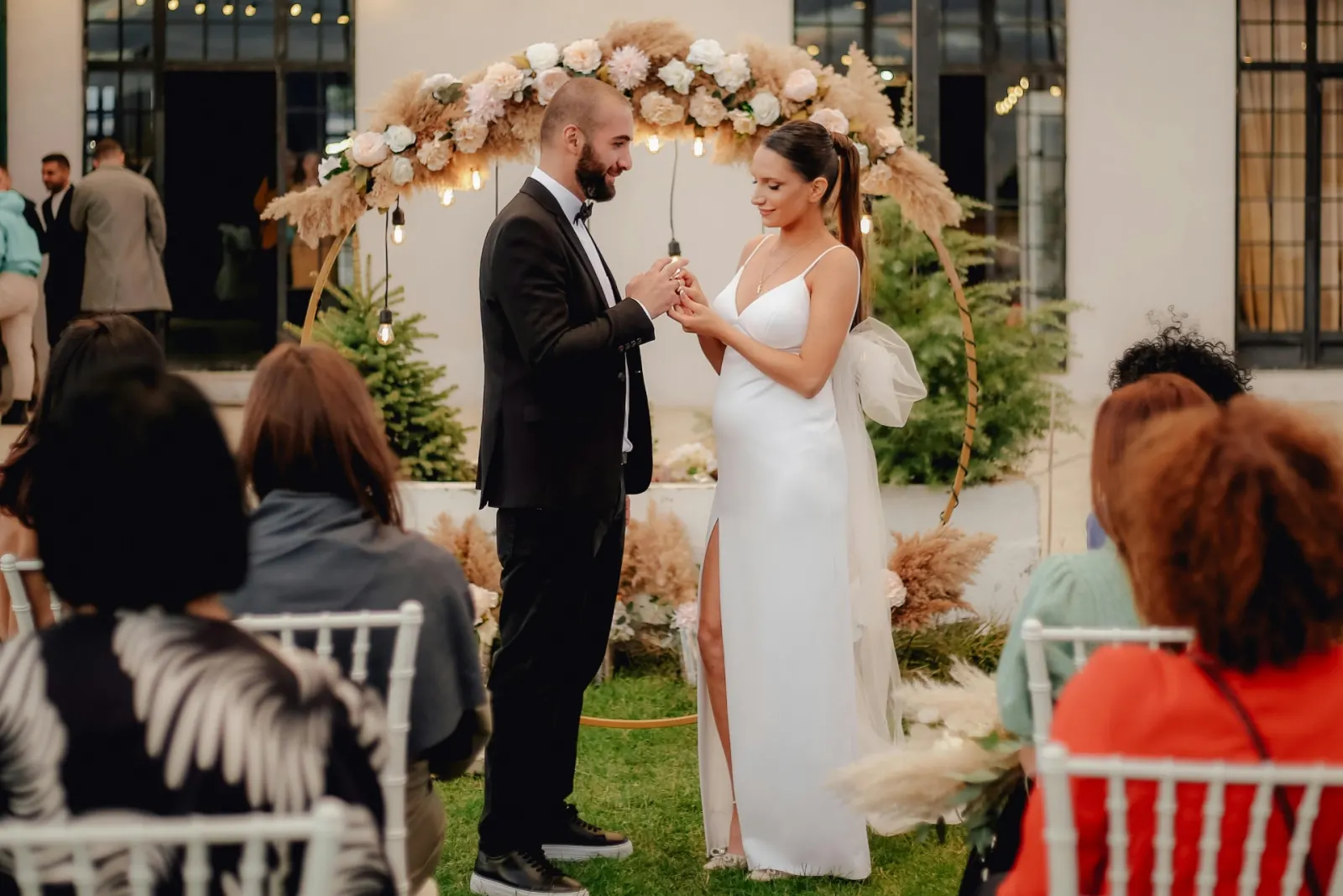 Bride and groom exchanging rings under a pampas grass and rose floral arch surrounded by guests