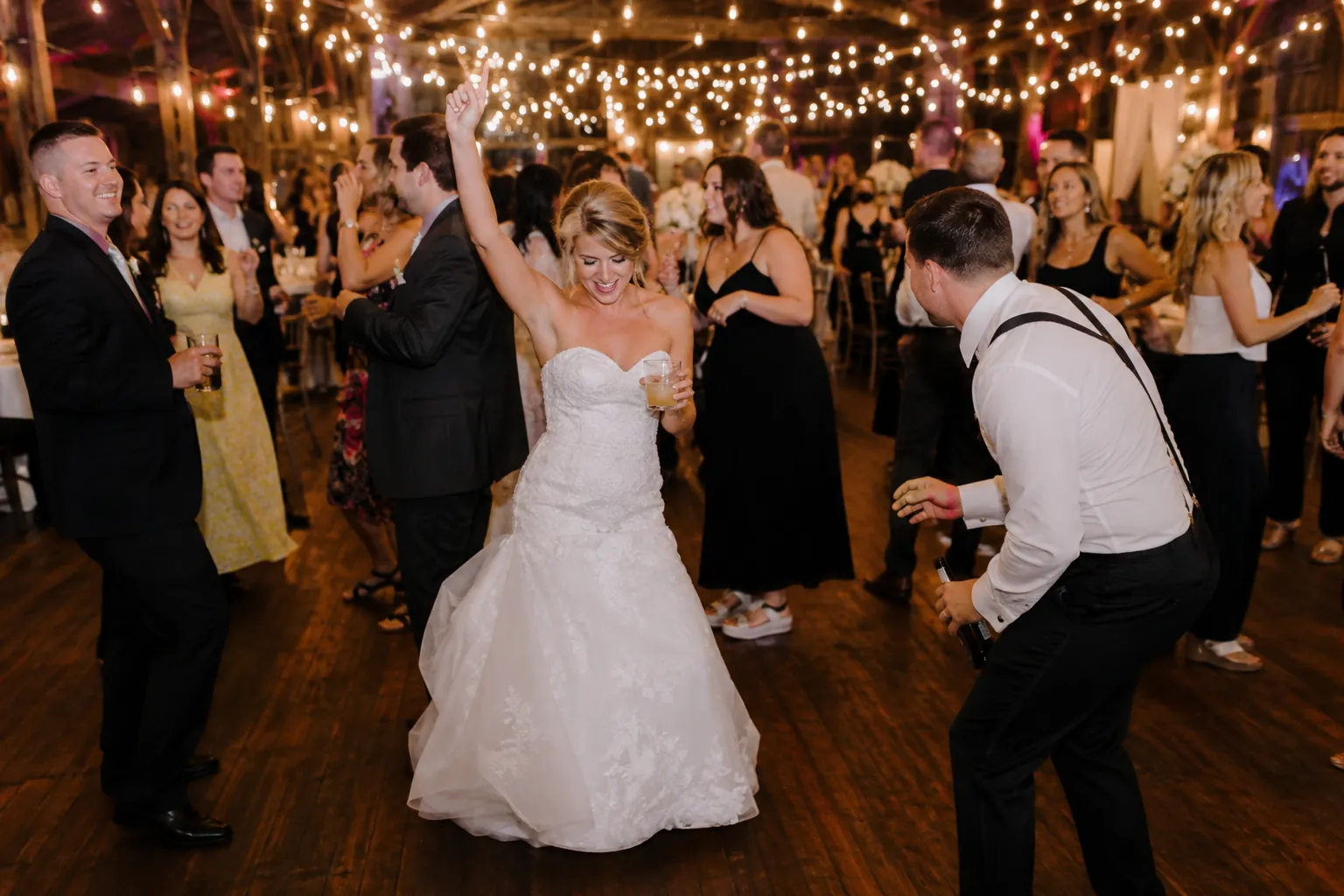 Bride dancing with her arm raised and drink in hand surrounded by guests under string lights on a wooden dance floor