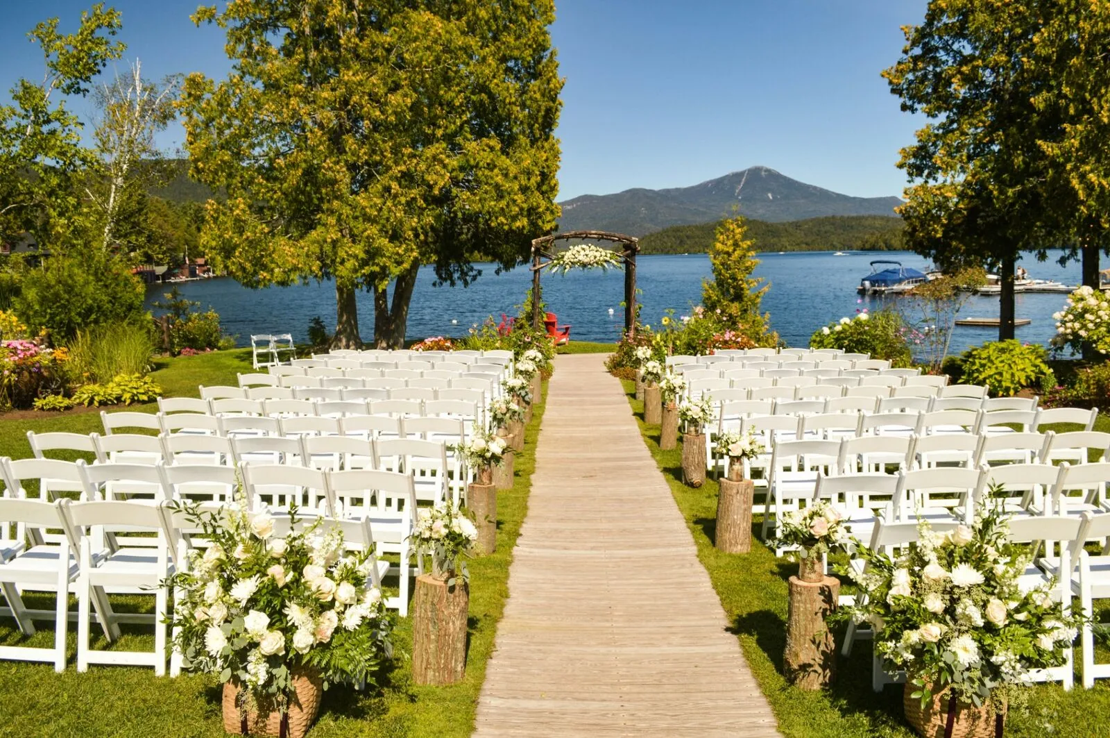 Outdoor lakeside wedding ceremony aisle with white chairs, floral arrangements, and a wooden arch
