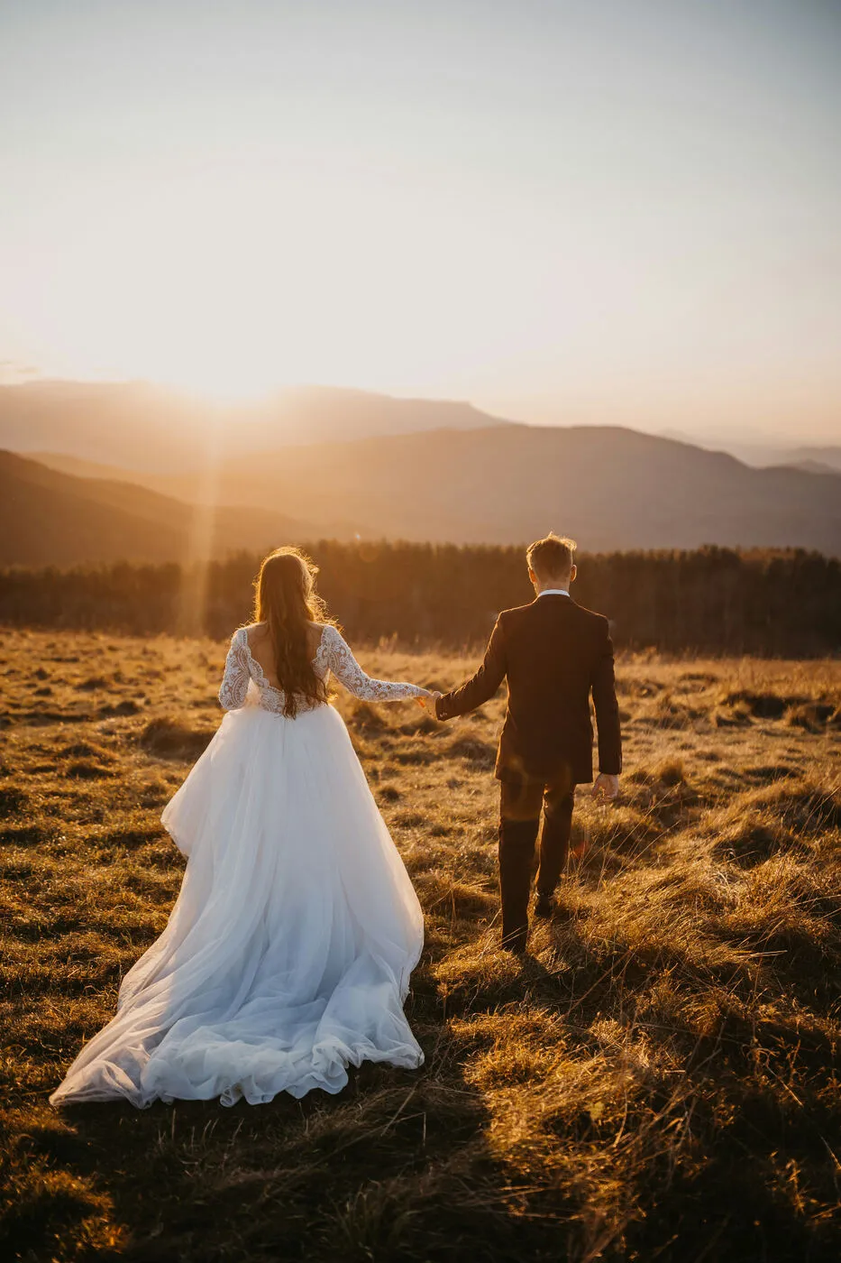 Bride and groom holding hands walking through a golden field at sunset in the mountains