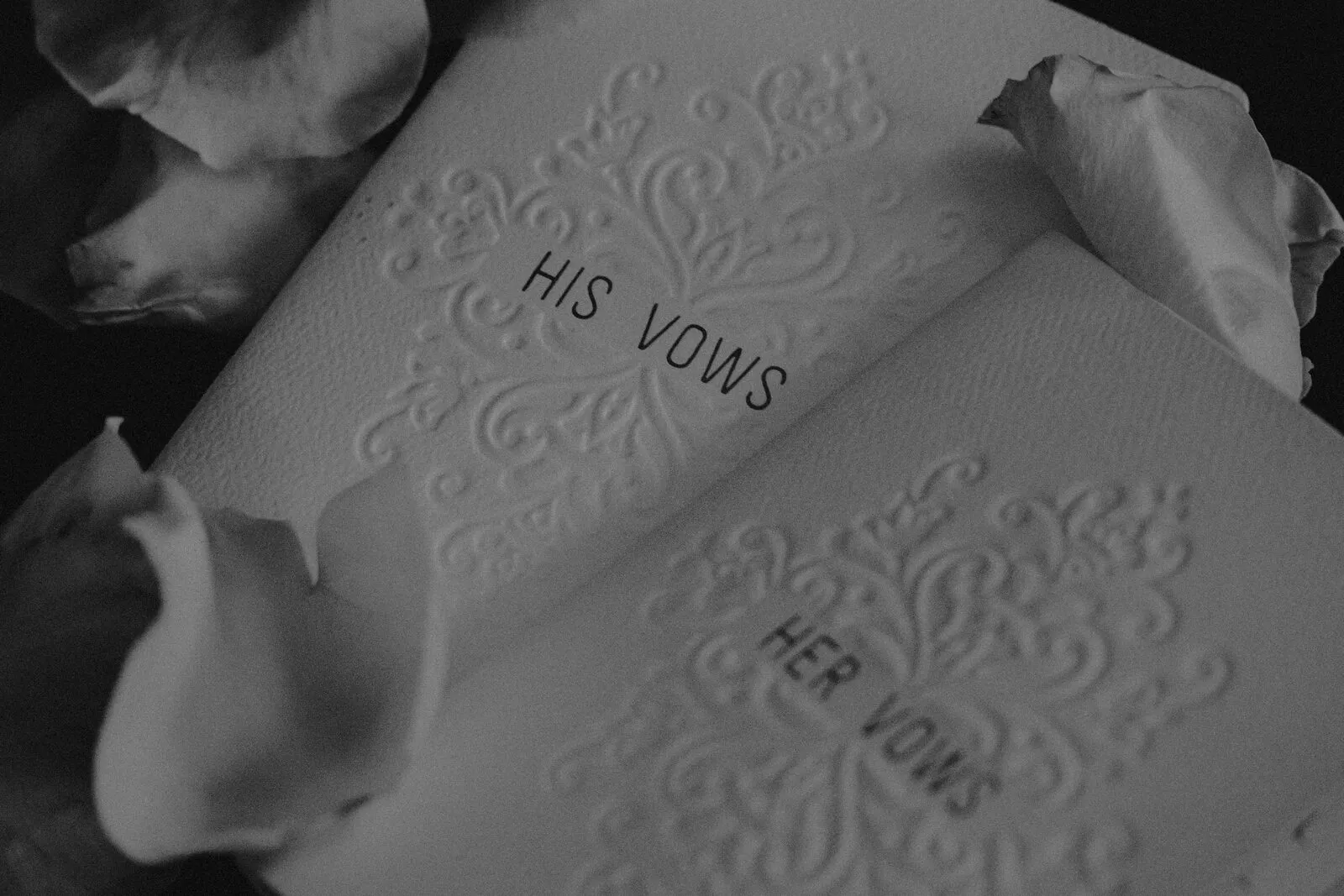 Black and white close-up of embossed His Vows and Her Vows booklets resting on white rose petals
